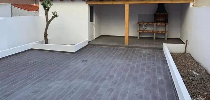 Pátio depois da obra, com pavimento imitação madeira, oliveira num canteiro branco e pérgola ao fundo.